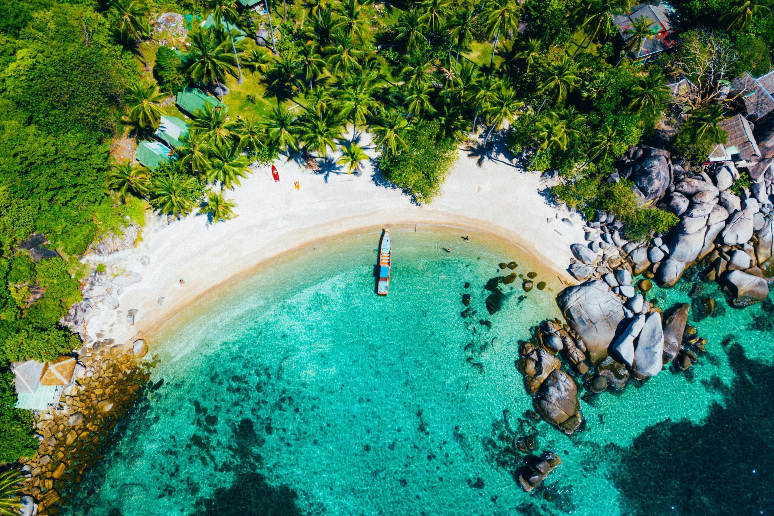 Aerial view of wooden boat docked at white sand beach with turquoise blue water, green trees and rocks